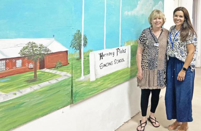 THE SHAPE OF PRAISE A hymnal for the harmony plains school (ab0ve) features the shaped notes of the solfege system. Below, Kathy Taylor (left) poses with daughter Barbara Lowrance in front of a mural depicting the front of the Harmony Plains Singing School in Cone, Texas, just over the Floyd County line in northern Crosby County. The school took over a former rural public school building and still gathers there every summer for instruction, singing, and fellowship. | LIZ ADAMS PHOTOS